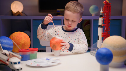 Blond boy paints planet solar system Mars with colorful paints sitting home table in evening,...
