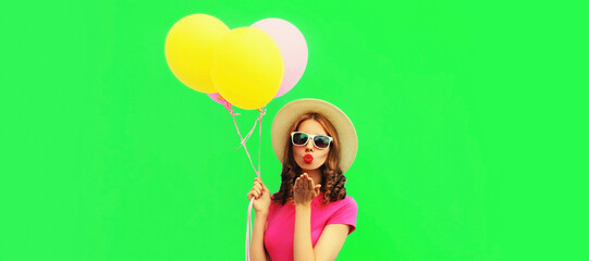 Young woman with colorful balloons blowing her red lips sending sweet air kiss wearing summer straw hat on green background