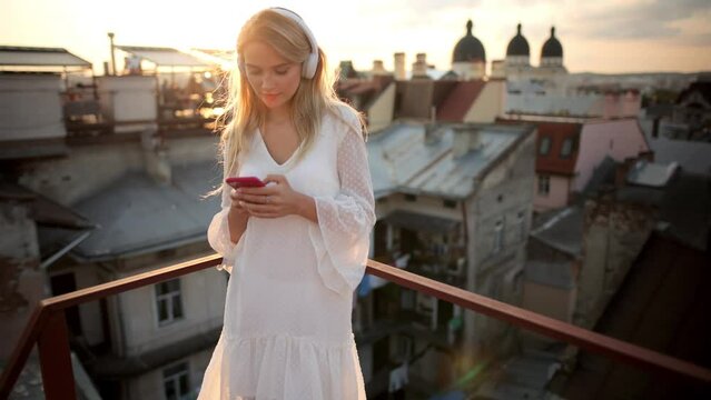 Happy young girl in earphones rest on Roof Terrace at Sunset. Peaceful blonde woman in modern wireless headphones relax on rooftop listening to music. Enjoy good quality sound. Stress free concept