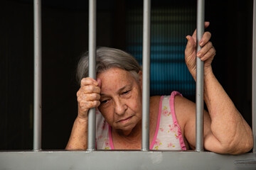 Mulher idosa com cara triste atrás das grades no portão.