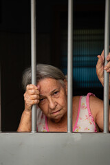 Mulher idosa com cara triste atrás das grades no portão.