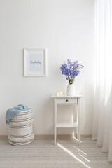Home interior decor. White bedside table, vase with bouquete of violet bell flowers, candles, wicker basket, frame with text SIMPLICITY. Light modern stylish room.