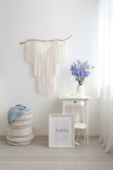Home interior decor. White bedside table, vase with bouquete of violet bell flowers, candles, wicker basket, frame with text SIMPLICITY, macrame wall hanging decoration. Light modern stylish room.