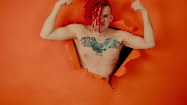 Shirtless man with red dreadlocks and with bird tattoo on chest, showing biceps and looking at camera through hole in orange wall in studio