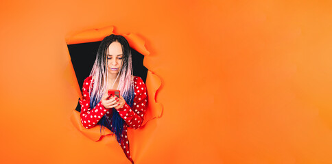 Bright woman browsing mobile phone and sticking out of hole of orange background. Impressive female with multicolored hairstyle using smartphone.