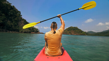 Strong man with thin plait hairstyle rows pink plastic kayak putting up paddle on sea against hills with wild jungles and blue sky. Young guy is sailing on kayak in ocean. Traveling to tropics.