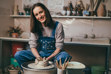 Smiling female potter working on pottery wheel while sitting in her workshop