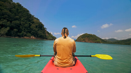 Young man with sunglasses and hat rows pink plastic canoe along sea against green hilly islands...