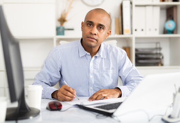 Adult businessman working in office with laptop, sitting at desk, looking at camera