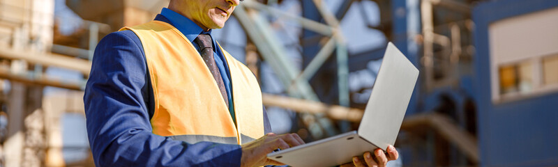 Matured man in work vest standing on territory of production plant and using modern notebook