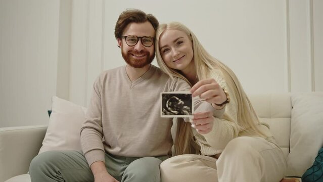 Cheerful millennial caucasian wife hugging her husband and holding ultrasound picture of their future baby while sitting on a sofa in their living room. High quality 4k footage
