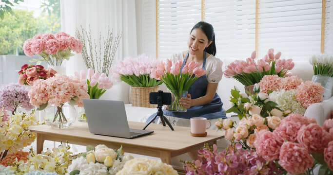 Asia girl vlogger influencer or SME owner people smile work on home video camera selfie shoot filming for live sell show happy talk on mobile VoIP app. Remote sale product at modern florist gift shop.