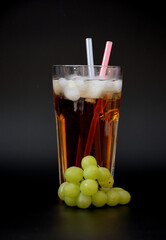 A tall glass of grape juice with ice and straws on a black background, next to a sprig of ripe fruits.