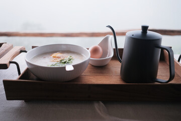 The winter traditional Thai breakfast is set in wooden tray on table, tasty morning meal, hot gruel, white cup of boiled rice with fried garlic and coriander, egg in a spoon, and a kettle of coffee.