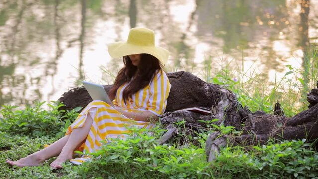 Asian woman leaning on driftwood log near lake at park, looking at tablet screen, reading message email, chatting with clients online, working remotely. Working and relaxing atmosphere in the park.