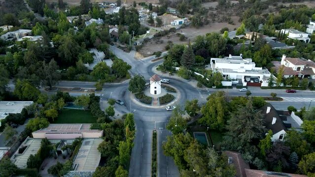 Aerial dolly in lowering on Leonidas Montes windmill in roundabout with vehicles commuting surrounded by trees, Lo Barnechea, Santiago, Chile