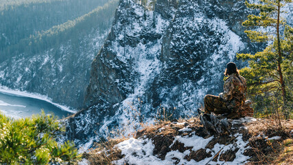 A young man in camouflage clothes sits on a log on a sunny slope of a rock against the backdrop of mountains covered with snow.