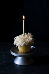 A vanilla cupcake topped with icing, coconut shavings and a lit candle.