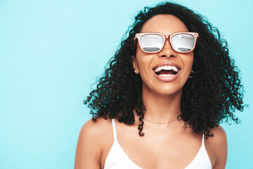 Fototapeta premium Beautiful black woman with afro curls hairstyle. Smiling model dressed in white summer dress. Sexy carefree female posing near blue wall in studio. Tanned and cheerful. In sunglasses. Closeup