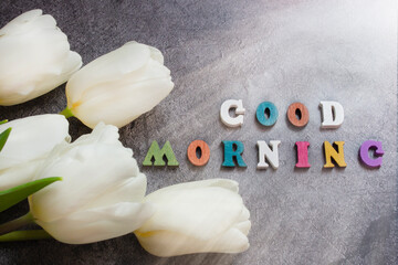 The inscription in letters Good morning is a phrase made of wooden letters on a dark concrete background with tulips flowers and sunlight.