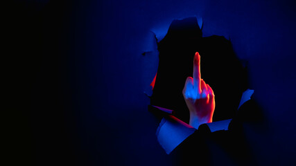 Fuck you. Provocative gesture. Red blue neon light female hand showing middle finger obscene sign inside breakthrough paper hole isolated on dark night copy space banner.