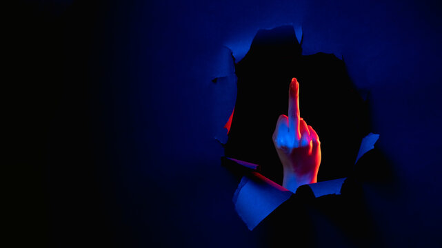 Fuck you. Provocative gesture. Red blue neon light female hand showing middle finger obscene sign inside breakthrough paper hole isolated on dark night copy space banner.