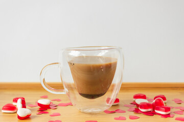 Glass double bottom cup of coffee on the wooden table with red paper hearts and candies.