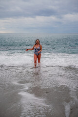 Fototapeta premium A plump woman in a bathing suit enters the water during the surf. Alone on the beach, Gray sky in the clouds, swimming in winter.