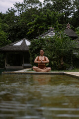 Young woman with body positive appearance practicing yoga alone on deck by the pool in tropical island of Bali, Indonesia. Sport, fitness, healthy lifestyle concept.