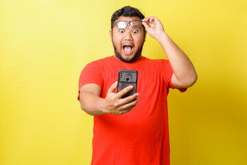 Fat asian man with surprised expression opening mouth while raising his glasses and looking to cell...