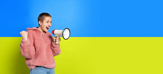 Portrait of young short- haired girl shouting in megaphone isolated over blue and yellow ukrainian...