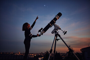 Girl with astronomical telescope stargazing under twilight sky. - obrazy, fototapety, plakaty
