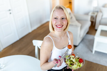 Portrait of a happy playful girl eating fresh salad from a bowl in her kitchen. Beautiful fit woman eating healthy salad after fitness workout