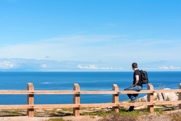 Fototapeta premium Man with backpack sitting on the fence at the end of earth watching on bright blue lake. Hiking or travel concept. Exploring the great outdoors. Beauty of nature
