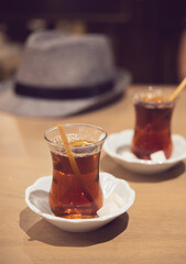 Two glasses with aroma traditional Turkish tea with sugar on white saucers close up on a wooden table in a cafe on the background of a hat. Istanbul, Turkey