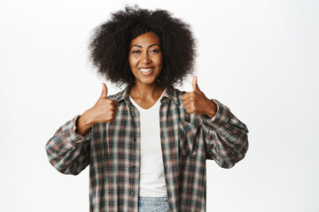Portrait of millennial african american woman, showing thumbs up, like and approve, praising smth, makes compliment, say yes, standing pleased against white background with happy smile
