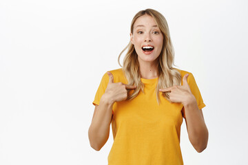 Obraz premium Enthusiastic young woman pointing fingers at herself, showing her, self-promoting, talking about personal achievement, standing over white background