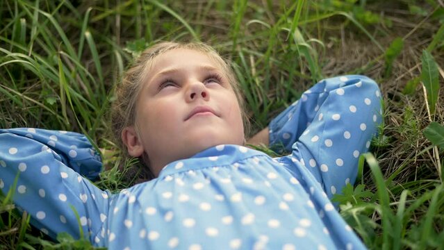 Happy little girl lies on green grass. The child is dream on the grass in park. Happy face of baby. Little girl looks at sky. Smile of baby. Girl is dream lies on grass. Happy baby face look up to sky