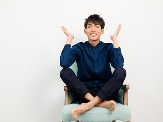 Portrait of handsome Chinese young man with curly black hair in blue shirt sitting in armchair and posing against white wall background. Smiling with arms open, full length front view studio shot