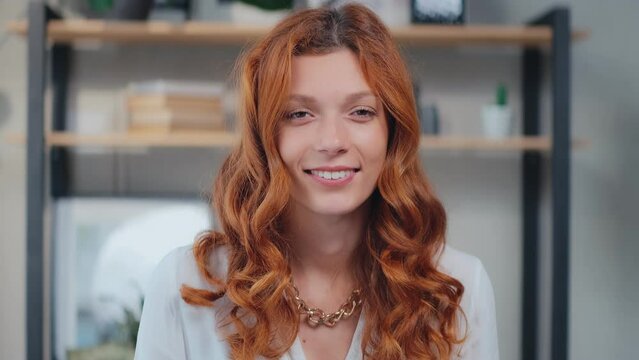 Young beautiful redhead Caucasian woman looking at camera cutely blinking and smiling listens to interlocutor during video call standing on background home interior. Headshot screen view