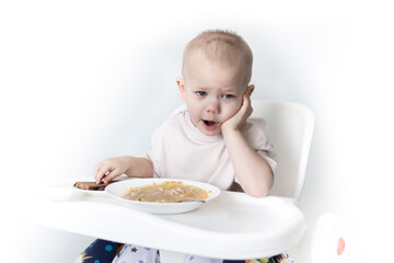 A little boy eats soup with bread on his own in a highchair against the background of a white wall.