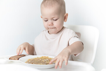 A little boy eats soup with bread on his own in a highchair against the background of a white wall.