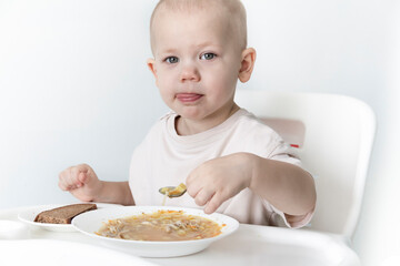 A little boy eats soup with bread on his own in a highchair against the background of a white wall.