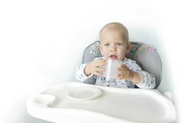 A child in a child seat in pajamas with stars drinks healthy yogurt against the background of a white wall.