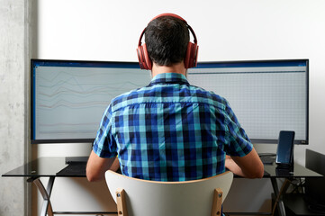 Back view of unrecognizable male employee in headphones reading graphs on computer screens while...