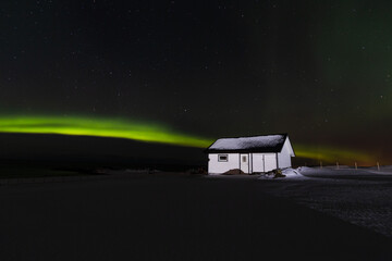 Mały skandynawski dom w nocy oświetlony piękną zorzą polarną, zielona zorza polarna przecina ciemne nocne niebo nad Lofotami w Norwegii