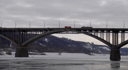 Obraz na płótnie Canvas Landscape with a span of a large arched bridge and a frozen river. A tram and a bus have met on the bridge and cannot separate. A tram with an empty passenger compartment is moving over a beautiful br