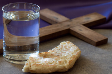 Lent season concept. Water, bread and cross on background of violet fabric