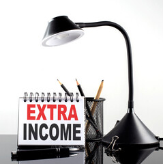 EXTRA INCOME text on notebook with pen and table lamp on the black background