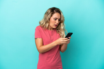 Young Brazilian woman isolated on blue background looking at the camera while using the mobile with surprised expression